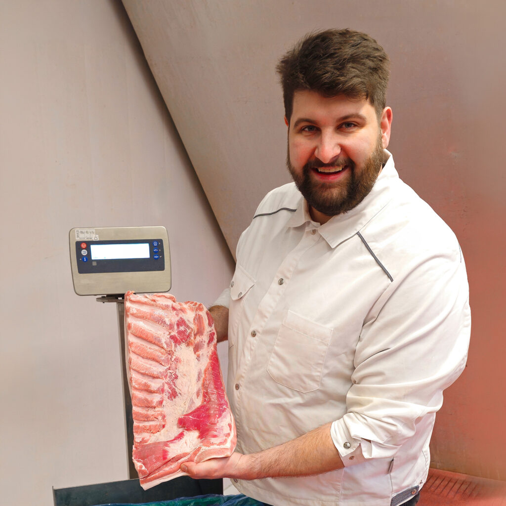 Person hält ein großes Stück rohes Schweinefleisch neben einer digitalen Waage in der Produktion.