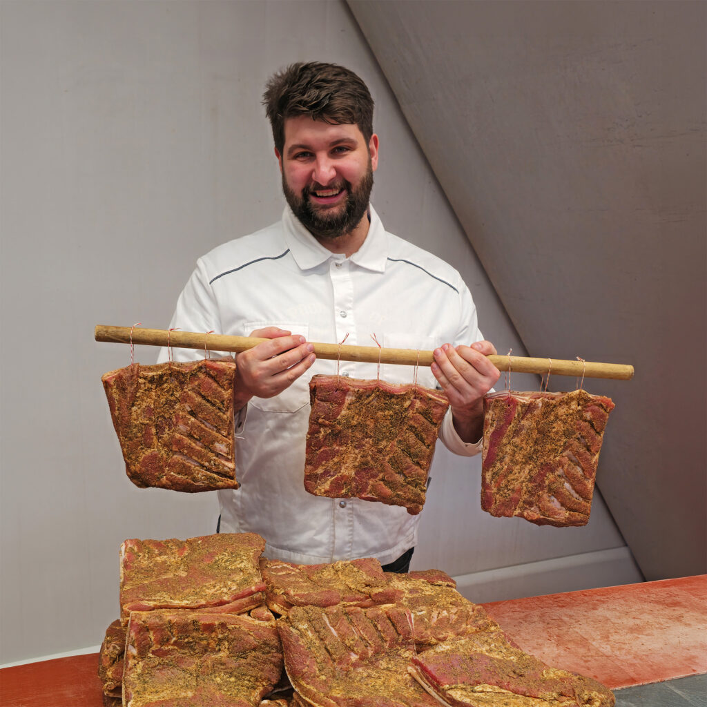 Person hält mehrere Stücke Buure Speck an einer Holzstange, weitere Speckstücke liegen auf dem Tisch.