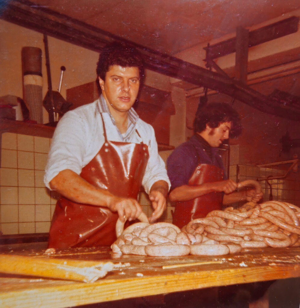Zwei Metzger mit roten Lederschürzen formen frische Würste von Hand auf einem Holzarbeitstisc in einer traditionellen Metzgerei – authentisches Handwerk aus der Schweiz.