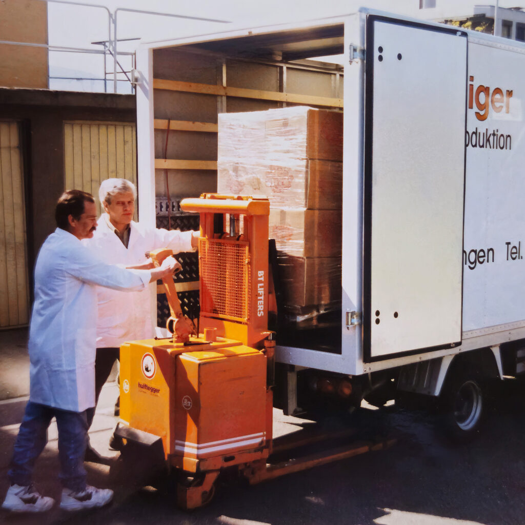 Zwei Mitarbeiter in weissen Kitteln beladen mit einem Elektrohubwagen einen Lieferwagen mit pallettierter Ware – zuverlässige Produktion und Lieferung von Speckmeister Luthiger.