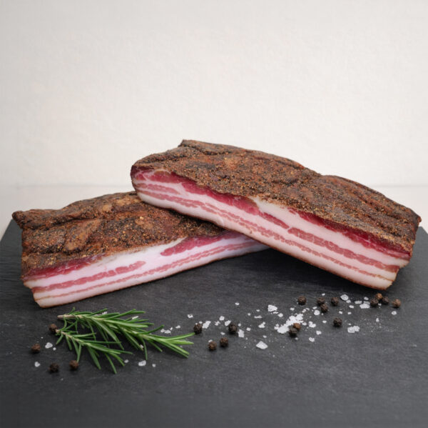 Zwei dicke Scheiben Buure Speck mit Pfefferkruste auf dunkler Steinplatte, umgeben von grobem Salz, Pfefferkörnern und einem Zweig Rosmarin.