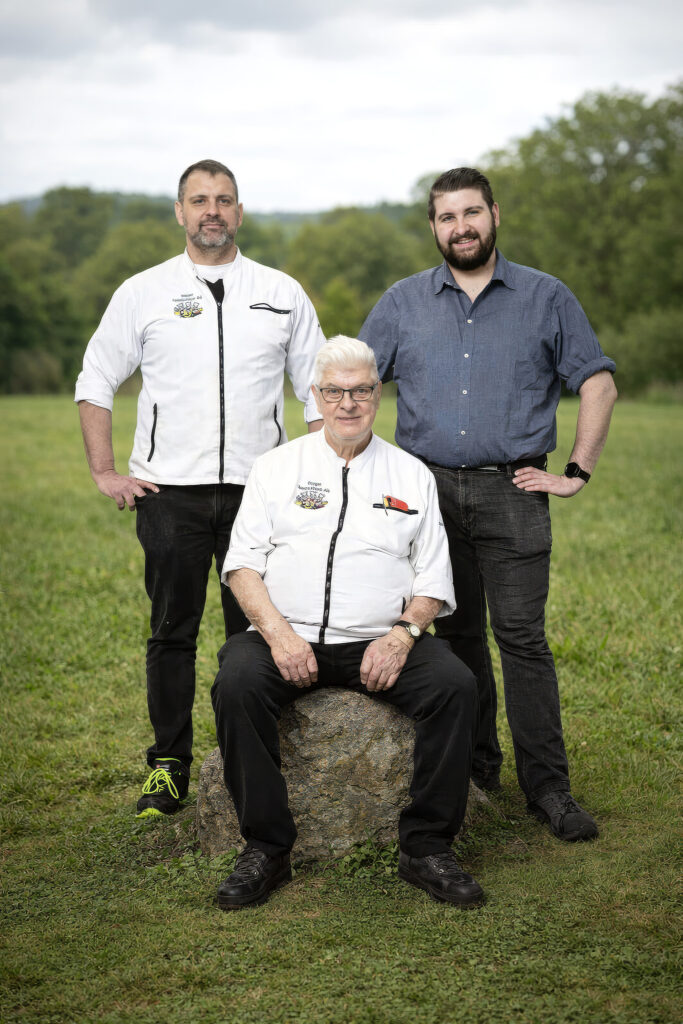 Drei Metzgerprofis im Freien; zwei in Kochjacken und einer im Hemd als Teamfoto der Speckmeister‑Familie.