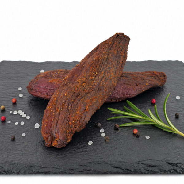 Getrocknetes, gewürztes Trockenfleisch auf Schieferplatte – rustikale Präsentation Zwei getrocknete, gewürzte Fleischstücke liegen auf einer Schieferplatte, umgeben von grobem Salz, bunten Pfefferkörnern und einem Rosmarinzweig.