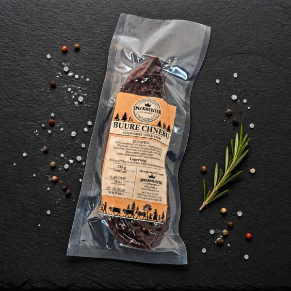 Buure Chnebu von Speckmeister – naturgereift und geräuchert Vakuumverpackter Buure Chnebu von Speckmeister liegt auf einer Schieferplatte, umgeben von grobem Salz, bunten Pfefferkörnern und einem Rosmarinzweig.