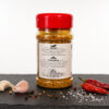 Gewürzdose mit Chili‑Knoblauch‑Mix auf dunkler Fläche, umgeben von Knoblauch, Pfefferkörnern, Salz und getrockneten Chilis.
