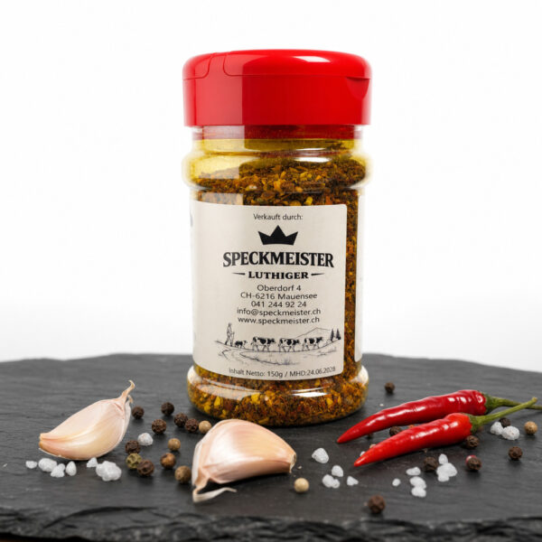 Speckmeister Luthiger Gewürzdose auf dunkler Fläche, umgeben von Chili, Knoblauch, Pfefferkörnern und grobem Salz.