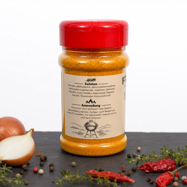 Gewürzdose mit gelb‑orangener Würzmischung auf dunkler Fläche, umgeben von Zwiebeln, Chili, Pfefferkörnern und Kräutern.