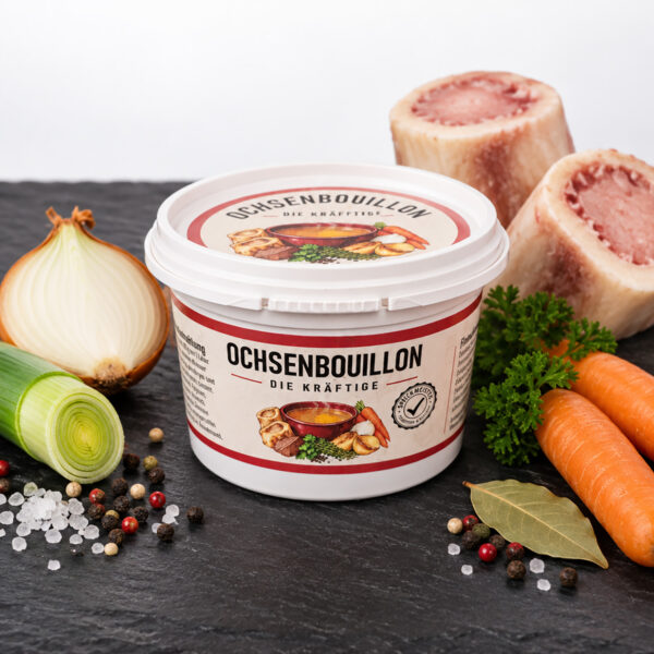 Ochsenbouillon 150 g in kleiner Dose mit weissem Deckel, Frontansicht mit Etikette «Die Kräftige», auf Schieferplatte mit Markknochen, Zwiebel, Lauch, Karotten, Lorbeer, Petersilie, Pfeffer und Meersalz
